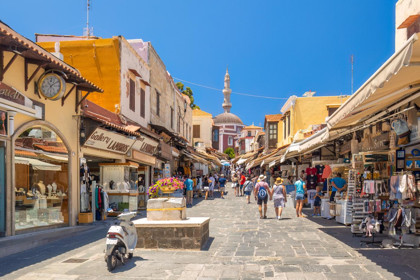 Tourists walking along a shopping street in Rhodes Old Town with traditional shops and Suleiman Mosque, Greece